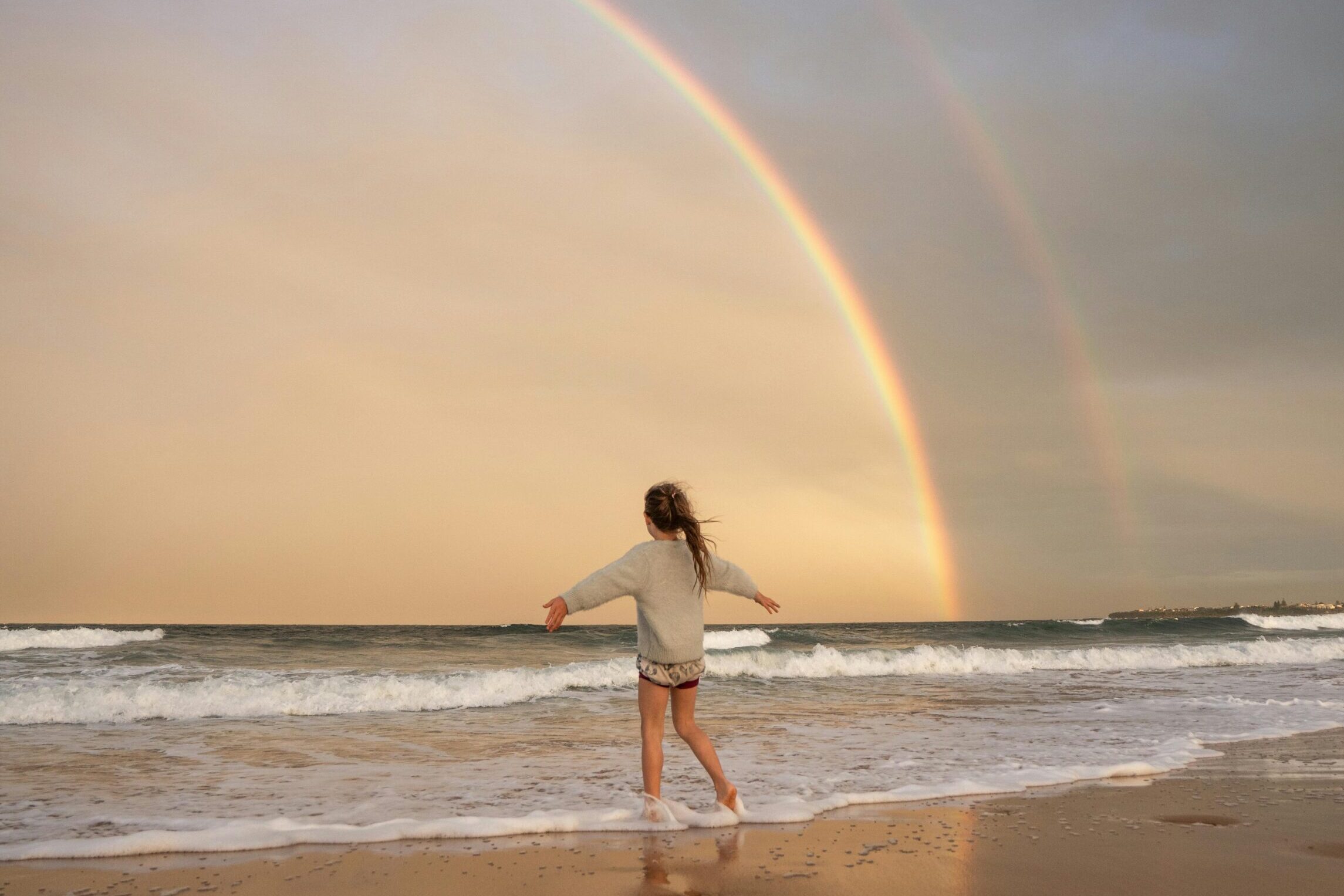 A woman joyfully runs along a beach beneath a vibrant rainbow arching over the sea.